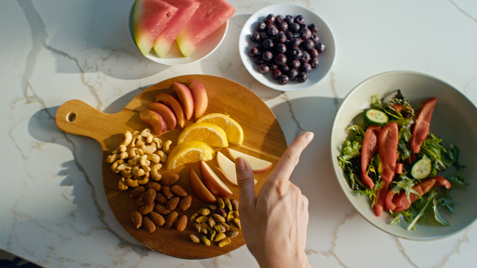 Hand zeigt auf bunte Auswahl an Früchten, Nüssen und Salat – gesunde Ernährung als Teil des Longevity Lebensstils Merz Aesthetics