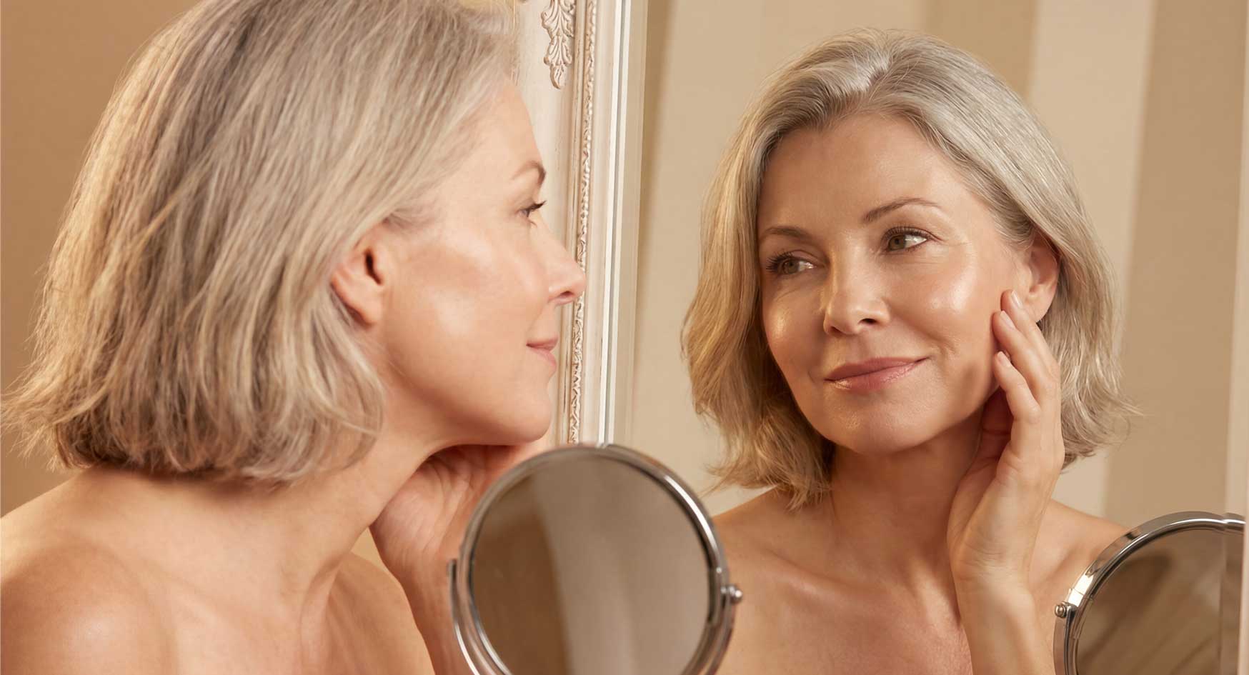 Frau betrachtet zufrieden ihr Gesicht im Spiegel | Natürlich aussehende Ergebnisse Anti-Aging Wechseljahre | Merz Aesthetics