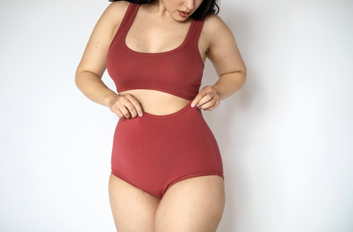 Frau in roter Unterwäsche berührt ihren Bauch, Nahaufnahme der Körpermitte vor hellem Hintergrund.