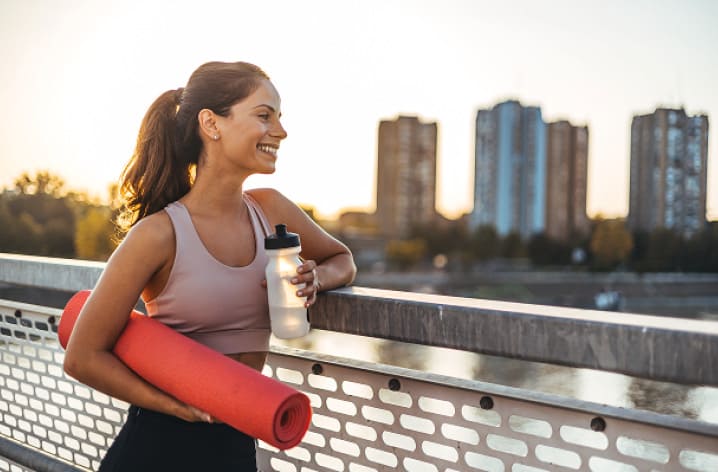 Frau in Sportkleidung mit Yogamatte und Trinkflasche lehnt an Brückengeländer, Skyline im Sonnenuntergang im Hintergrund.