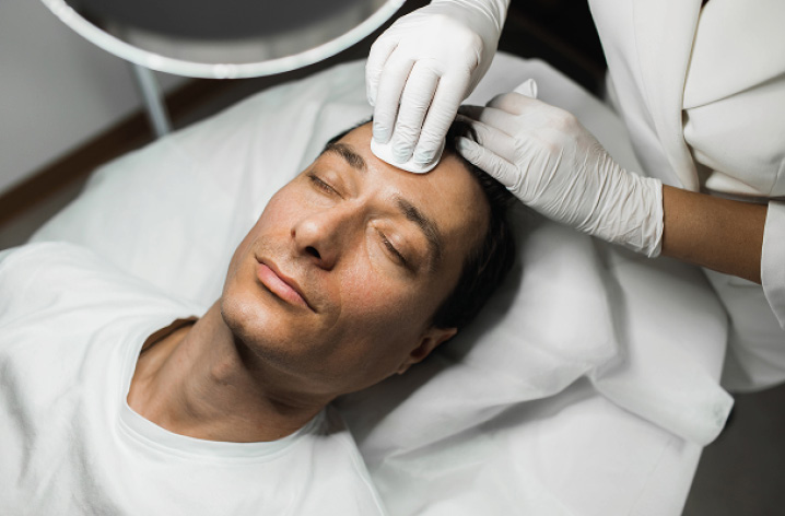Mann liegt entspannt mit geschlossenen Augen, Ärztin mit Handschuhen berührt seine Stirn, Behandlungsraum im Hintergrund