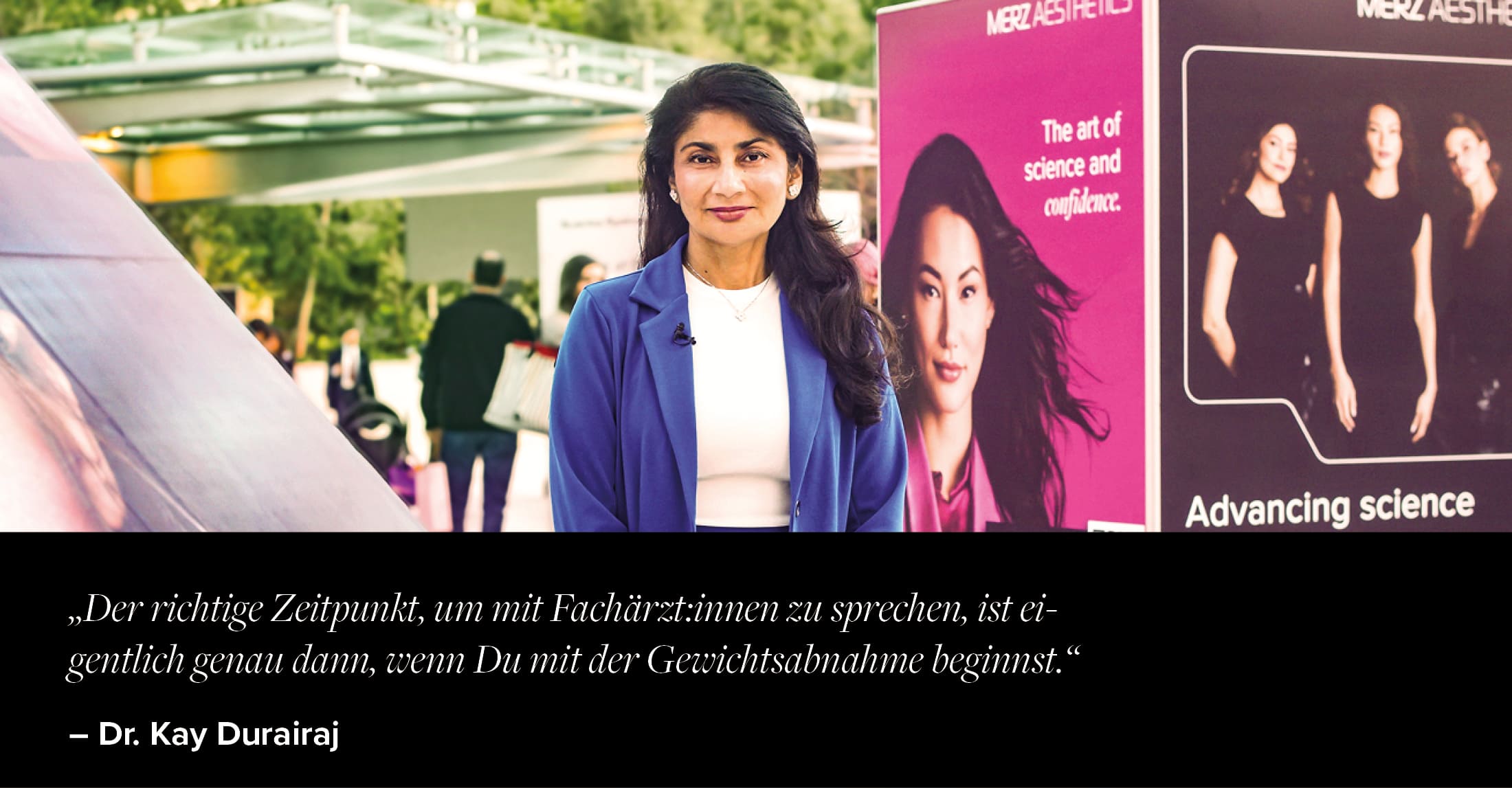 Dr. Kay Druairaj im blauen Blazer steht vor Kongressständen von Merz Aesthetics® mit Werbebannern, Kongressgelände im Hintergrund 