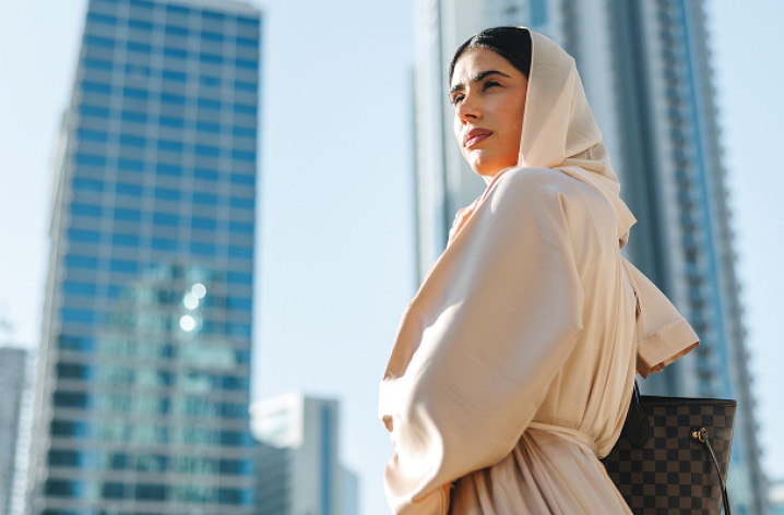 Frau mit beigem Kopftuch und Handtasche blickt zur Seite, Wolkenkratzer und Sonnenlicht im Hintergrund einer Stadt