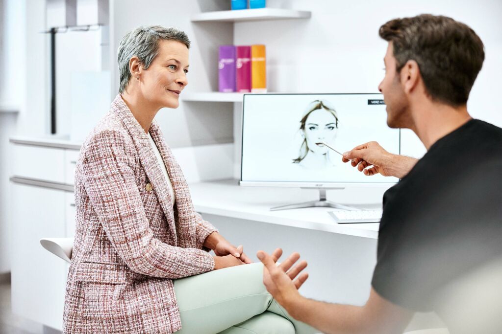 Arzt von Merz Aesthetics® zeigt einer Patientin am PC Behandlungsmöglichkeiten.
